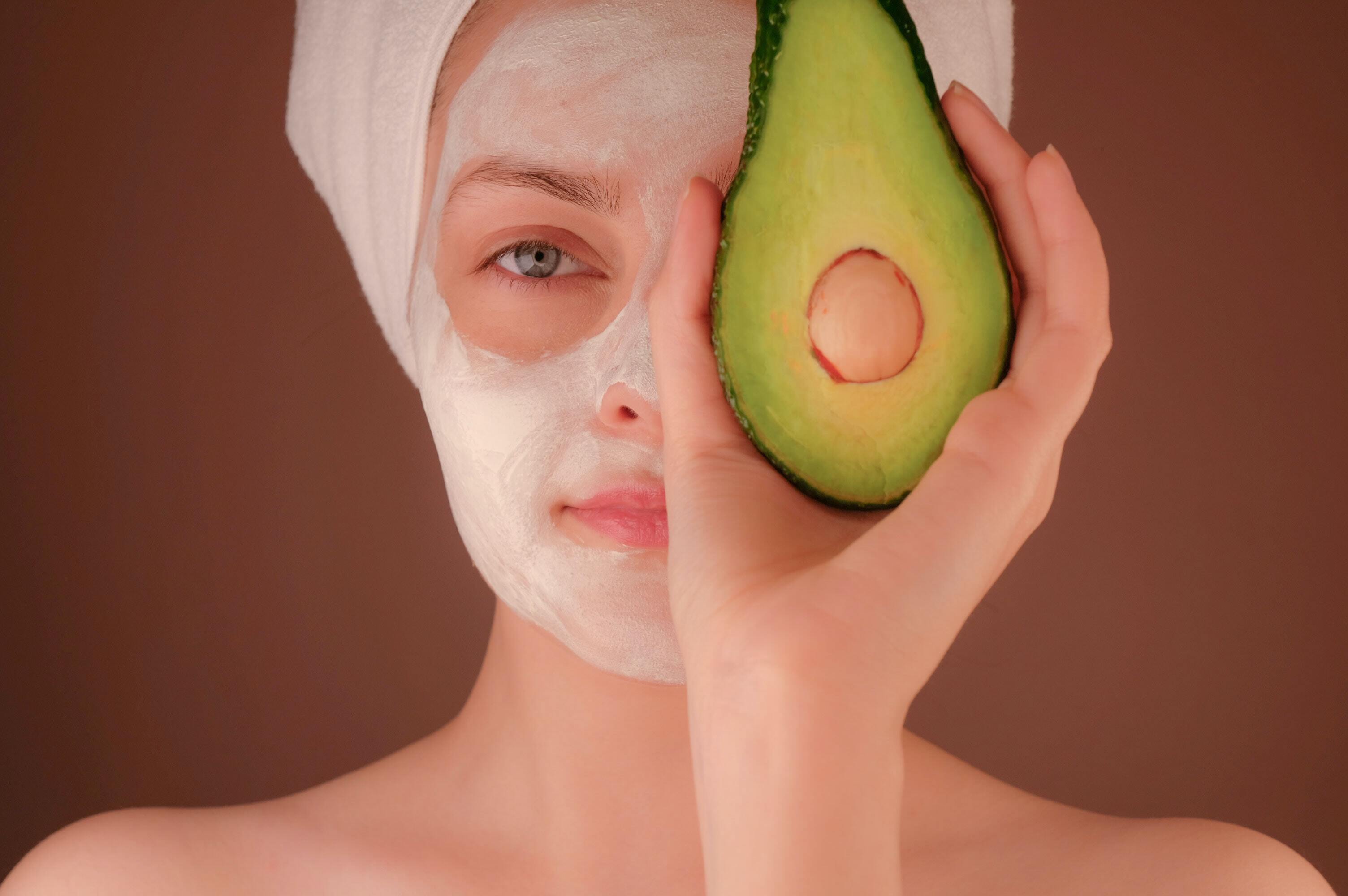 Woman holding avocado to her face, with a face mask on.